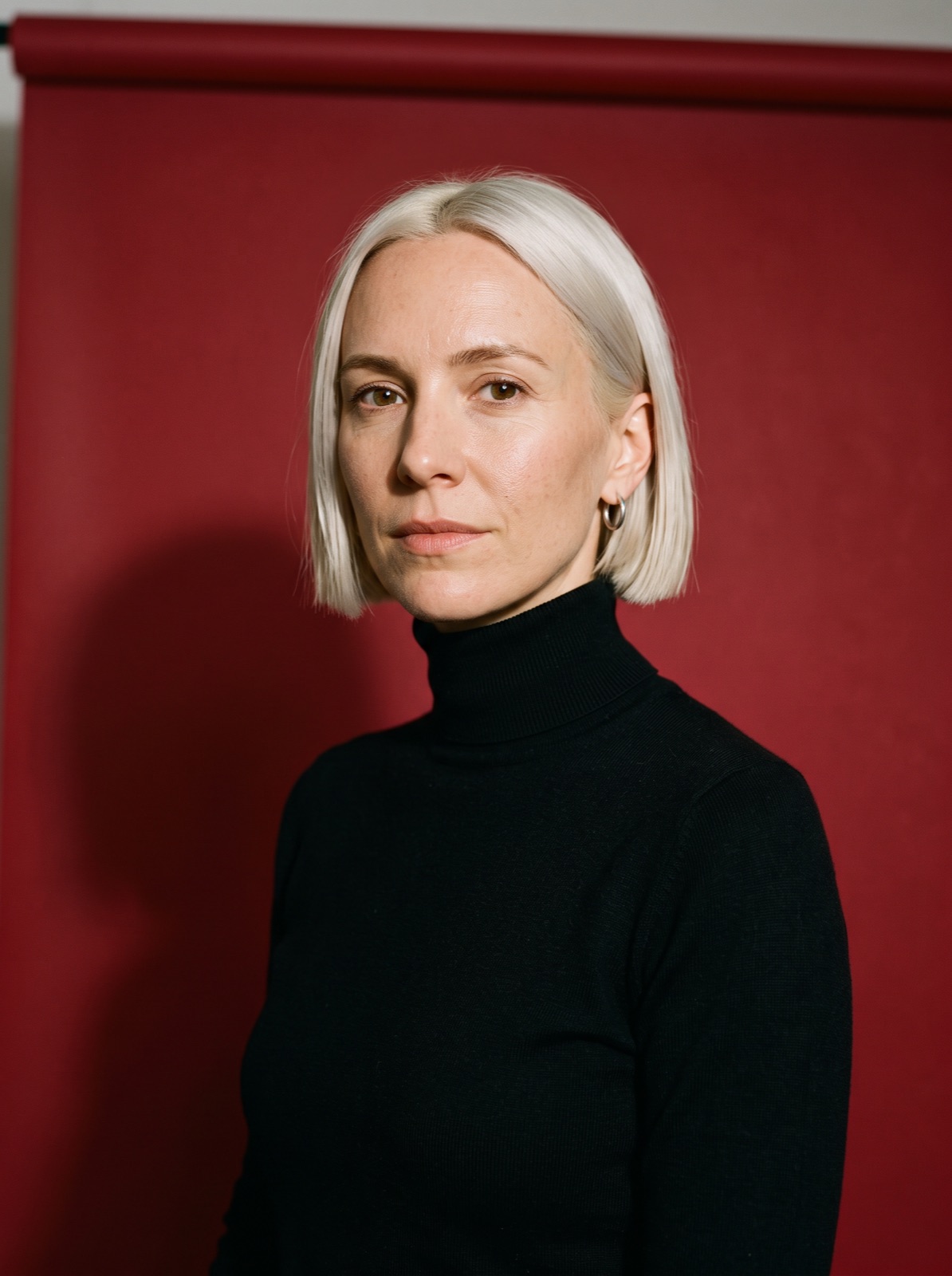 Wende Kroon, platinum bob and black roll-neck, in front of a red paper backdrop